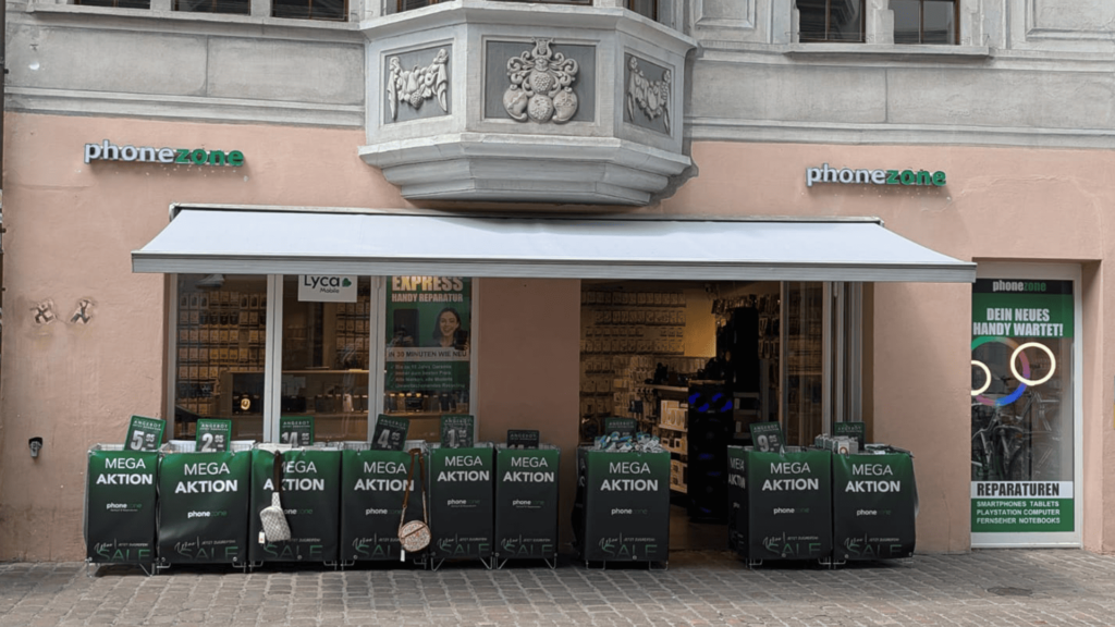 Ladenfront des Mobiltelefongeschäfts phonezone in Schaffhausen. Die Fassade zeigt das Firmenlogo über einer ausgedehnten Markise, Plakate für Express-Handyreparaturen sowie zahlreiche grüne Körbe mit der Aufschrift „MEGA AKTION“ im Aussenbereich.