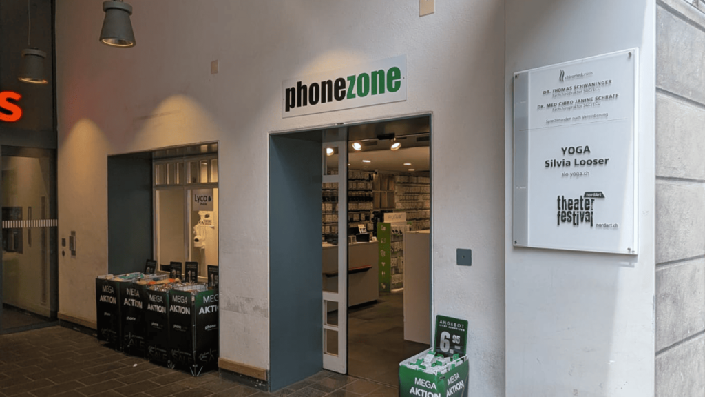 Eingangsbereich von phonezone in einer Passage oder einem Bogengang. Neben der offenen Tür stehen Aktionskörbe und an der Wand befindet sich ein weisses Praxisschild für Chiropraktik, Yoga und ein Theaterfestival.